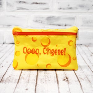 May include: Yellow zippered pouch with an orange zipper and the embroidered text "Oooo, Cheese!". The pouch has a cheese pattern with orange circles. The background is a white brick wall and a wooden surface.