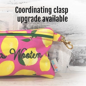 May include: Pink zippered pouch with a lemon print and the text "Wooten" embroidered on the front. The pouch has a gold metal clasp and a green zipper. The pouch is made by "PERSONally" and has a coordinating clasp upgrade available.