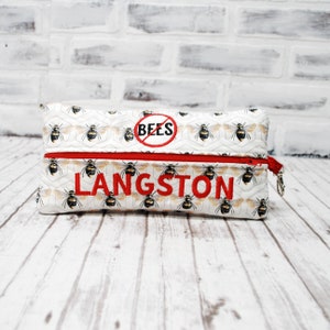 May include: White zippered pouch with a bee pattern and red zipper. The pouch has a red circle with a diagonal line through it and the word "BEES" inside. The name "LANGSTON" is embroidered in red on the front of the pouch.