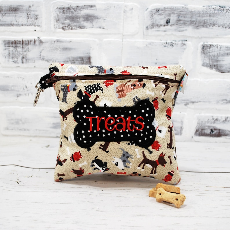 May include: A beige fabric treat pouch with a zipper closure and a black bone-shaped patch with white polka dots and the word "Treats" embroidered in red. The pouch is decorated with a pattern of cartoon dogs.