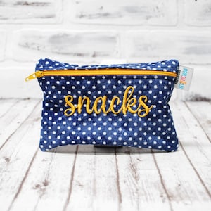 May include: A blue and white polka dot zippered pouch with the word "snacks" embroidered in yellow. The pouch is made of fabric and has a yellow zipper.