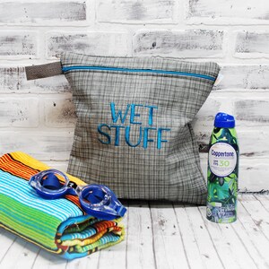 May include: A gray and white zippered pouch with the text "WET STUFF" embroidered in blue. The pouch is sitting on a white wooden surface next to a colorful beach towel, blue swimming goggles, and a green and blue sunscreen bottle with the brand name "Coppertone" and SPF 30.