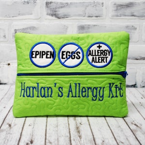 May include: Green zippered pouch with blue trim and embroidered text that reads "Harlan's Allergy Kit". Three round blue and white patches are embroidered on the pouch, reading "EPIPEN", "EGGS" with a diagonal line through it, and "ALLERGY ALERT" with a plus sign.