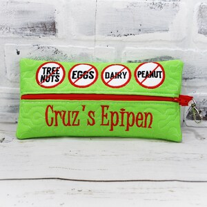 May include: Green zippered pouch with red zipper and red embroidered text "Cruz's EpiPen". Four red circles with white text are embroidered on the pouch: "Tree Nuts", "Eggs", "Dairy", and "Peanut".