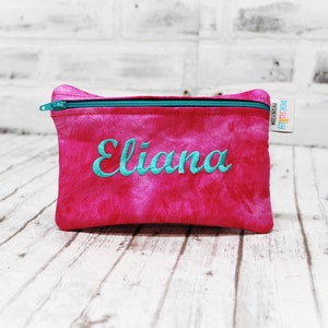 May include: Pink zippered pouch with a tie-dye pattern and the name "Eliana" embroidered in turquoise.