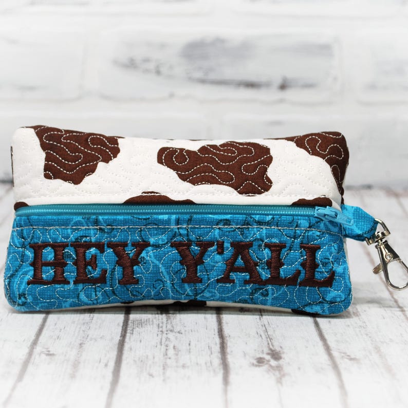 May include: A small zippered pouch with a cow print design and a blue fabric lining. The pouch has a zipper closure and a brown embroidered phrase that reads "Hey Y'all".
