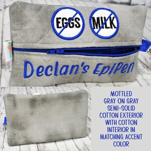 Op de afbeelding: Een gewatteerd grijs tasje met rits, voorzien van twee ronde geborduurde patches met blauwe randen, elk met een blauwe schuine streep over de woorden EGGS en MILK. De tekst Declan's EpiPen is in blauw garen onder de rits geborduurd.