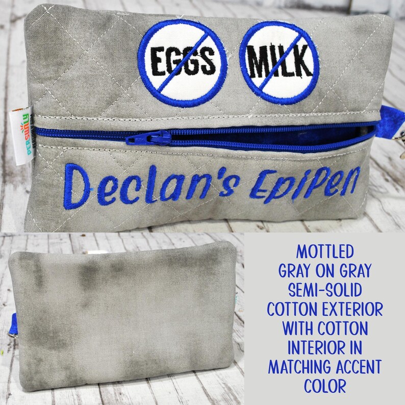 Op de afbeelding: Een gewatteerd grijs tasje met rits, voorzien van twee ronde geborduurde patches met blauwe randen, elk met een blauwe schuine streep over de woorden EGGS en MILK. De tekst Declan's EpiPen is in blauw garen onder de rits geborduurd.