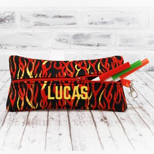 May include: Red and yellow flame patterned pencil case with a zipper closure. The case has a gold zipper pull and a gold name tag that reads "LUCAS".
