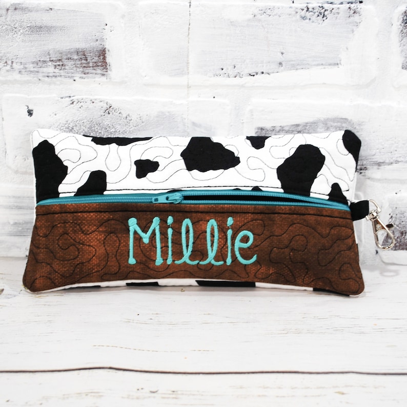 May include: A brown and black cow print zippered pouch with a brown faux leather bottom. The pouch has a turquoise zipper and a silver clip. The name "Millie" is embroidered on the front in turquoise thread.