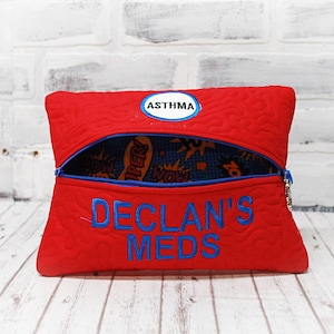 May include: Red quilted zippered pouch with blue trim and a blue embroidered label that says "ASTHMA". The pouch is open and the inside lining is blue with a comic book pattern. The pouch is embroidered with the text "DECLAN'S MEDS" in blue.