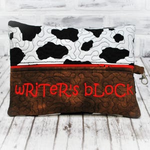 May include: A brown and black cow print zippered pouch with a red zipper and a brown fabric flap with the text "WRITER'S BLOCK" embroidered in red.