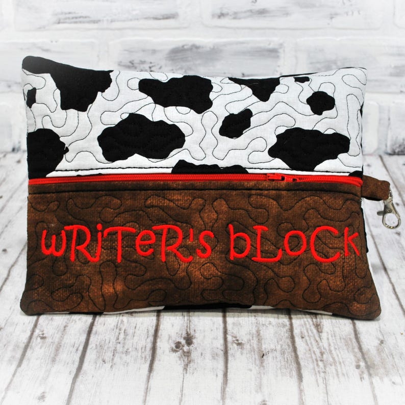 May include: A brown and black cow print zippered pouch with a red zipper and a brown fabric flap with the text "WRITER'S BLOCK" embroidered in red.