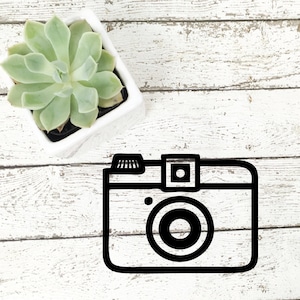 May include: Black and white illustration of a vintage camera on a white wood background.