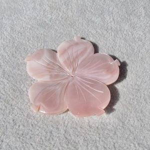 May include: A light pink, five-petal flower carving. The petals have detailed veining and a small hole in the center. The flower is likely made of a polished stone or shell material, and it is set against a textured, off-white background.