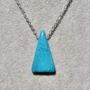 May include: A turquoise triangle pendant necklace with a silver chain. The pendant is a vibrant blue-green colour and is shaped like a pyramid. The chain is a delicate silver colour, and the necklace is displayed against a grey background.