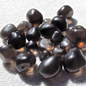 May include: A cluster of polished, dark brown gemstones with a smooth, reflective surface. The stones vary in size and shape, with a mix of rounded and irregular forms. They are arranged on a white, textured surface.