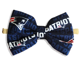May include: A navy blue bow tie with a repeating pattern of the word "PATRIOTS" and the team logo. The bow tie is attached to a tan elastic band.