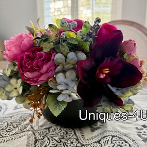 May include: A vibrant floral arrangement featuring a mix of pink, magenta, and burgundy flowers, accented with green foliage and gray-blue accents. The arrangement is in a black vase and sits on a patterned tablecloth. The text "Uniques-4U" is visible.