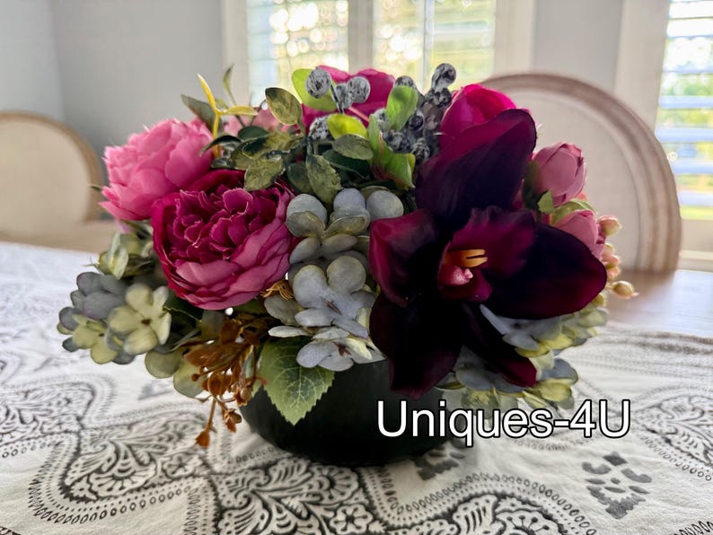 May include: A vibrant floral arrangement featuring a mix of pink, magenta, and burgundy flowers, accented with green foliage and gray-blue accents. The arrangement is in a black vase and sits on a patterned tablecloth. The text "Uniques-4U" is visible.