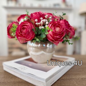 May include: A floral arrangement featuring vibrant red roses, white filler flowers, and green foliage in a pearlescent vase. The vase sits atop a book with the text "Uniques-4U". This decorative piece adds a touch of elegance.