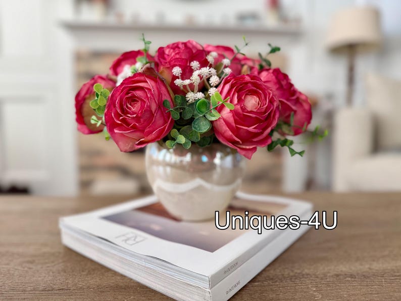 May include: A floral arrangement featuring vibrant red roses, white filler flowers, and green foliage in a pearlescent vase. The vase sits atop a book with the text "Uniques-4U". This decorative piece adds a touch of elegance.