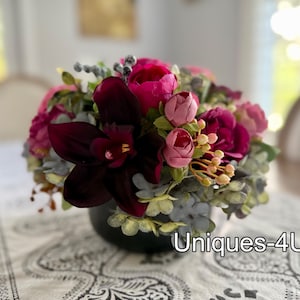 May include: A floral arrangement in a dark green vase. The arrangement features a mix of pink, fuchsia, and burgundy flowers, with accents of green and gray foliage. The text "Uniques-4U" is visible on the bottom right.