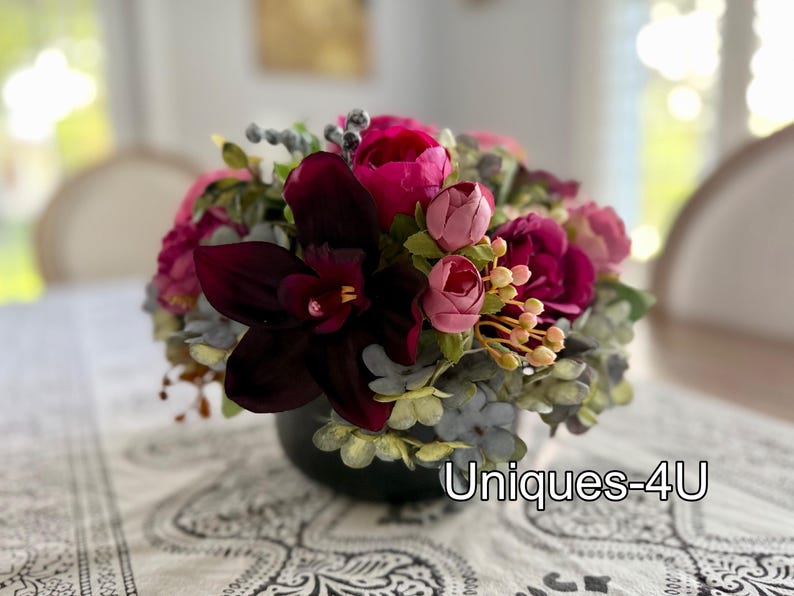 May include: A floral arrangement in a dark green vase. The arrangement features a mix of pink, fuchsia, and burgundy flowers, with accents of green and gray foliage. The text "Uniques-4U" is visible on the bottom right.