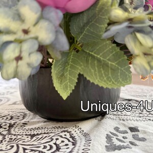 May include: A floral arrangement in a dark gray, round pot. The flowers are a mix of blue, pink, and green, with large leaves. The text "Uniques-4U" is visible on the pot.