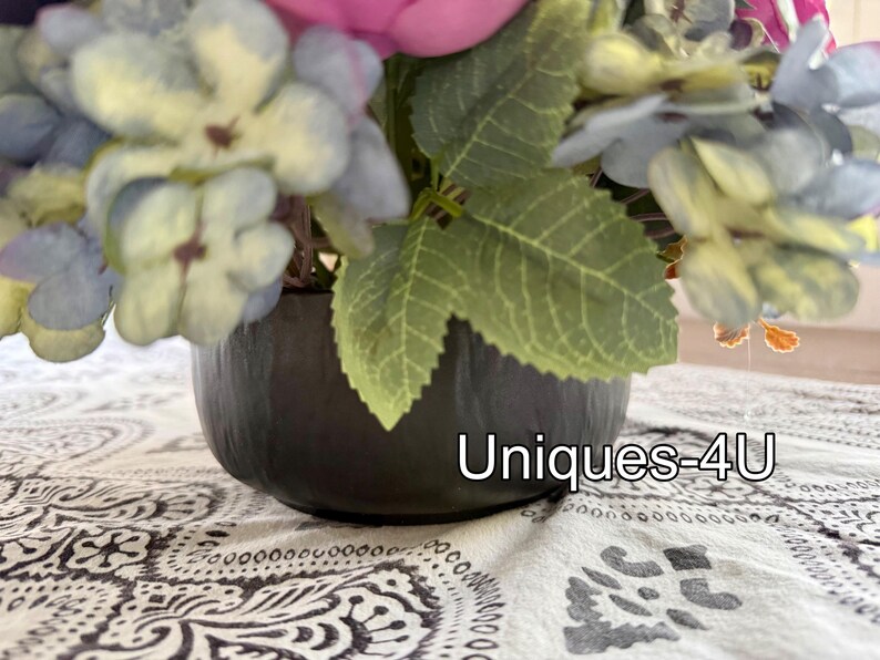May include: A floral arrangement in a dark gray, round pot. The flowers are a mix of blue, pink, and green, with large leaves. The text "Uniques-4U" is visible on the pot.