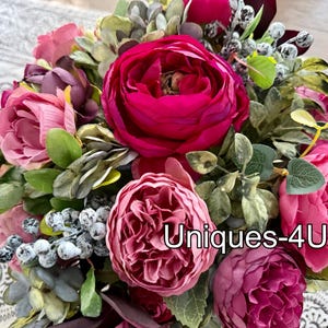 May include: A vibrant bouquet of artificial flowers, featuring large, lush blooms in shades of pink and magenta. Green foliage and berries add texture. The text "Uniques-4U" is visible.