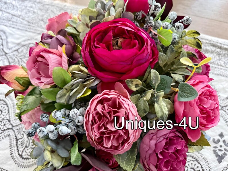 May include: A vibrant bouquet of artificial flowers, featuring large, lush blooms in shades of pink and magenta. Green foliage and berries add texture. The text "Uniques-4U" is visible.