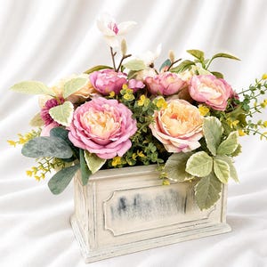 May include: A rectangular, distressed off-white wooden planter box filled with an arrangement of artificial flowers, including pink and peach roses, small yellow sprigs, and green foliage, topped with a single white magnolia blossom.