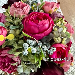 May include: A vibrant floral arrangement featuring large, deep pink peonies as the focal point, complemented by smaller pink and burgundy blooms. Green foliage and frosted berries add texture. The text "Uniques-4U" is visible.