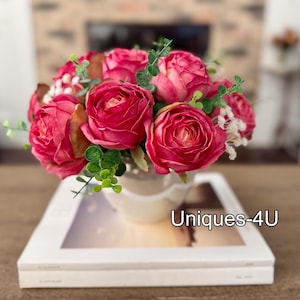 May include: A floral arrangement featuring vibrant pink roses, green foliage, and small white flowers in a cream-colored vase. The vase sits atop a white book with the text "Uniques-4U". The arrangement is a decorative item.