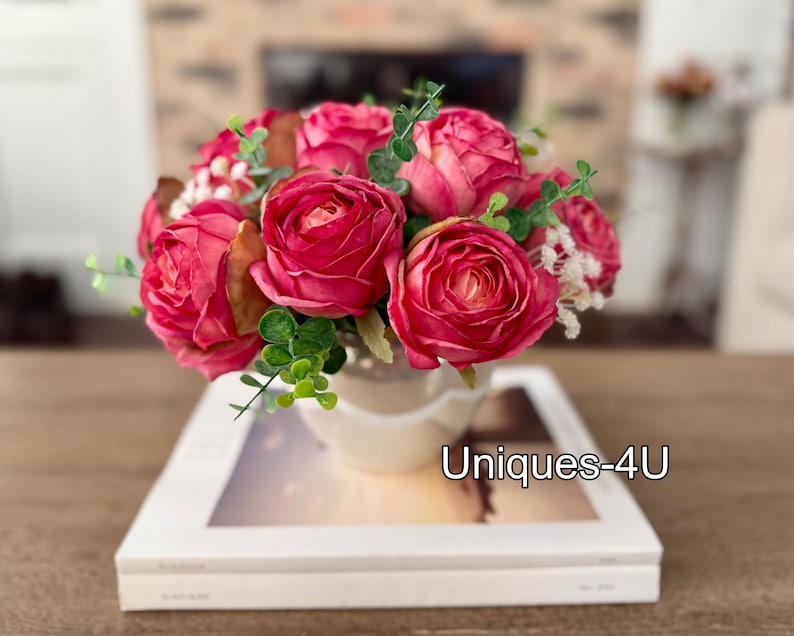 May include: A floral arrangement featuring vibrant pink roses, green foliage, and small white flowers in a cream-colored vase. The vase sits atop a white book with the text "Uniques-4U". The arrangement is a decorative item.