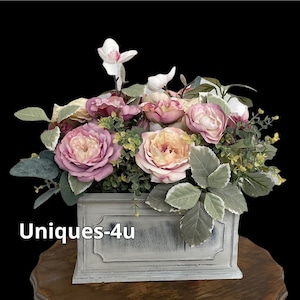 May include: A whitewashed wooden planter box filled with a floral arrangement of pink and cream roses, white orchids, and green foliage. The planter has a decorative design and the text "Uniques-4u" is visible on the front.