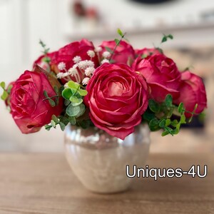 May include: A floral arrangement featuring vibrant red roses, small white flowers, and green foliage. The flowers are arranged in a pearlescent, round vase. The text "Uniques-4U" is visible on the vase.