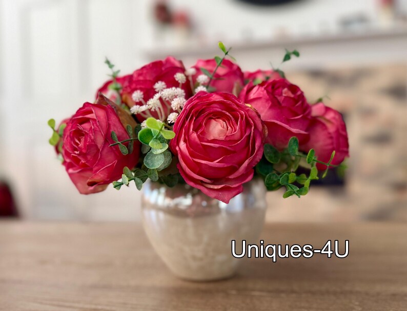 May include: A floral arrangement featuring vibrant red roses, small white flowers, and green foliage. The flowers are arranged in a pearlescent, round vase. The text "Uniques-4U" is visible on the vase.