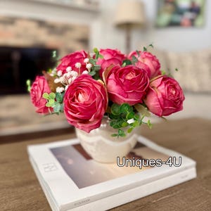 May include: A floral arrangement featuring vibrant pink roses and greenery in a cream-colored vase. The vase sits atop a stack of books with the text "RH OUTDOOR" and "RH INTERIORS". The text "Uniques-4U" is also visible.
