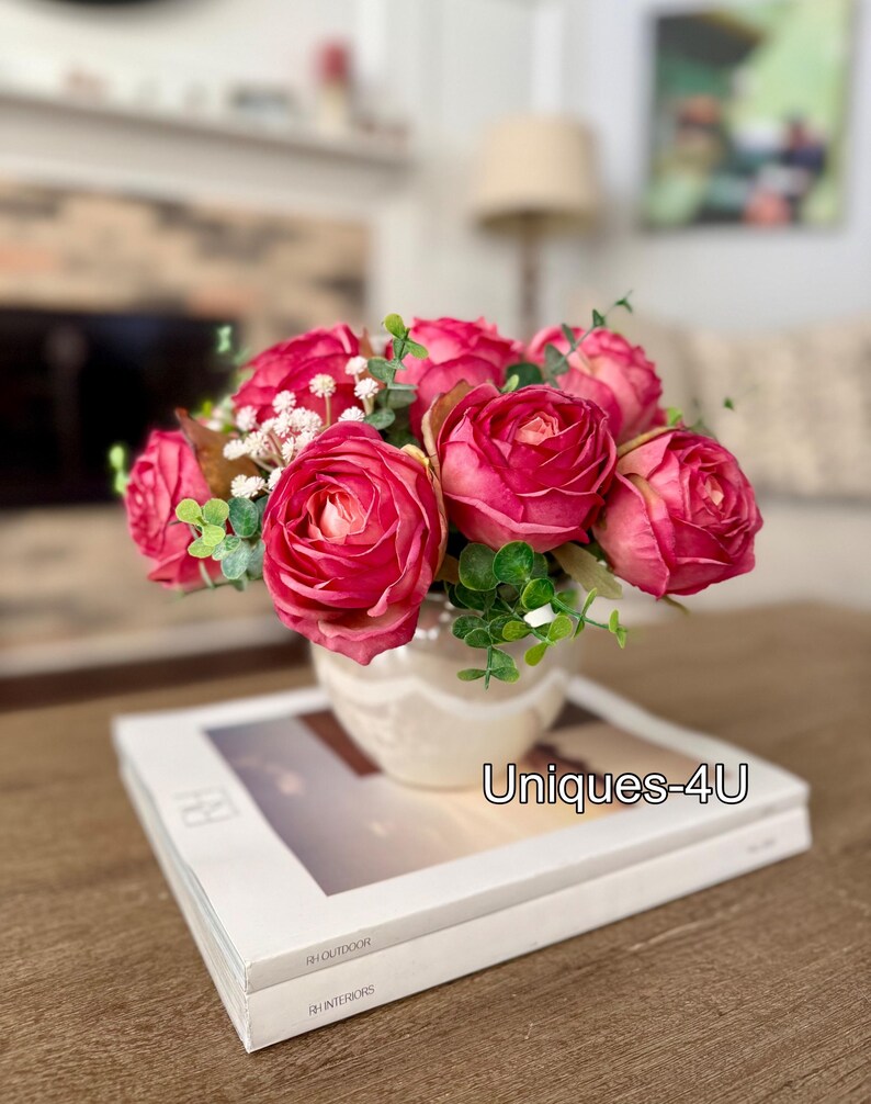 May include: A floral arrangement featuring vibrant pink roses and greenery in a cream-colored vase. The vase sits atop a stack of books with the text "RH OUTDOOR" and "RH INTERIORS". The text "Uniques-4U" is also visible.