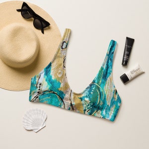 May include: A blue and gold patterned bikini top with a scoop neck. The top is on a white surface with a straw hat, sunglasses, and sunscreen.