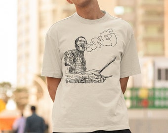May include: A cream-coloured t-shirt featuring a black and white sketch of a musician playing an instrument. The word "SONGS" is written above the musician. The person in the image is wearing a black cap and sunglasses and black trousers.