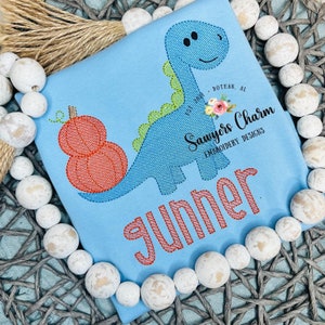 May include: A light blue t-shirt featuring a blue dinosaur holding a stack of orange pumpkins. The dinosaur has a green spine and a friendly face. The shirt also features the text "Gunner" in pink and "Sawyers Charm Embroidery Designs" in black.