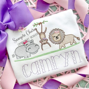 May include: White embroidered baby onesie with a hippopotamus, monkey, and lion design. The onesie has the name "Camryn" embroidered in purple below the animals.
