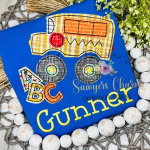 May include: A blue t-shirt featuring a school bus design with the name "Gunner" in yellow. The bus is yellow and orange, with black wheels and the letters "ABC" in colorful patterns. "Sawyers Charm Embroidery Designs" is also visible.