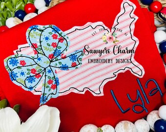 May include: Red t-shirt with an embroidered outline of the United States, a blue and white floral bow, and the text "Sawyers Charm Embroidery Designs" and "EST. 2021". The name "Lyla" is embroidered in blue below the outline of the United States.