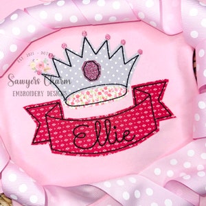 May include: Embroidered design of a silver crown with pink jewels on top of a red banner with the name "Ellie" in black lettering.