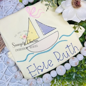 May include: A yellow and blue sailboat with a pink bow embroidered on a light yellow fabric. The name "Elsie Ruth" is embroidered in cursive below the sailboat.