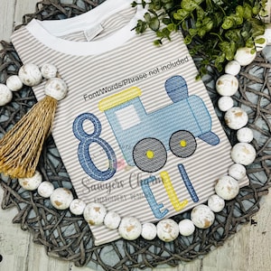May include: A light gray and white striped t-shirt with a blue train applique. The train has yellow and black accents, with the number 8 and the name "Eli" embroidered in blue and yellow. The shirt is on a woven mat with decorative beads.